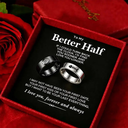A gift box containing two rings, one black and one silver, with a romantic message printed on the insert card. Red roses and a red fabric background surround the box.